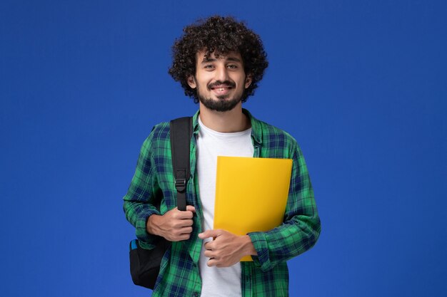 Vista frontal del estudiante en camisa a cuadros verde vistiendo mochila negra y sosteniendo archivos en la pared azul