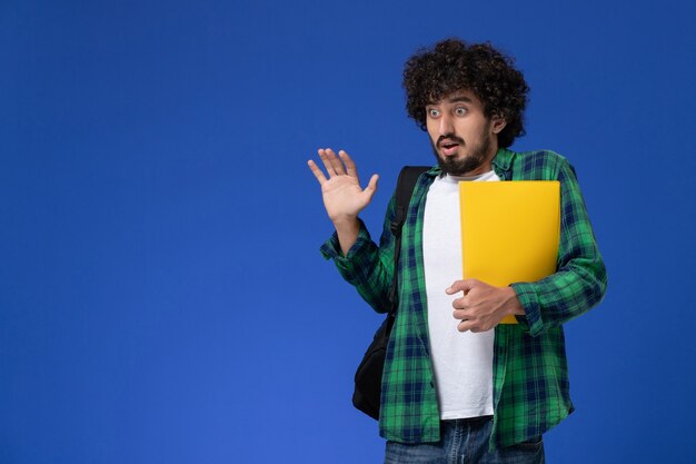 Vista frontal del estudiante en camisa a cuadros verde vistiendo mochila negra y sosteniendo archivos en la pared azul