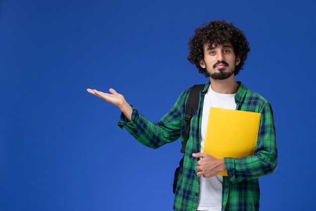 Vista frontal del estudiante en camisa a cuadros verde vistiendo mochila negra y sosteniendo archivos en la pared azul