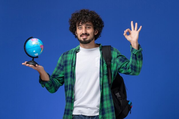 Vista frontal del estudiante en camisa a cuadros verde vistiendo mochila negra y sosteniendo un pequeño globo sonriendo en la pared azul