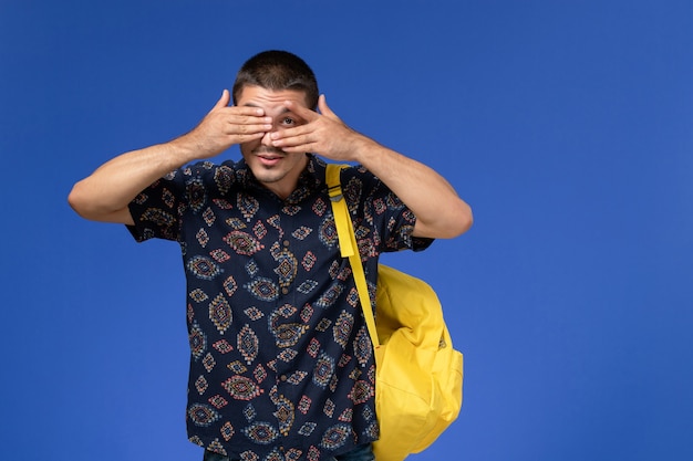 Vista frontal del estudiante en camisa oscura con mochila amarilla en la pared azul