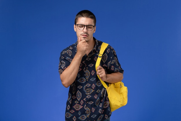 Vista frontal del estudiante en camisa oscura con mochila amarilla pensando en la pared azul claro