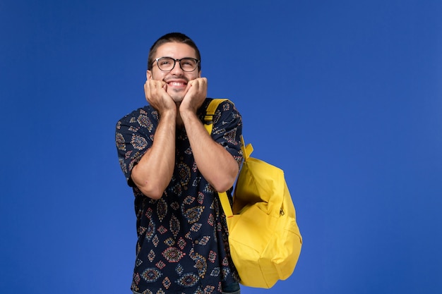 Vista frontal del estudiante en camisa oscura con mochila amarilla posando con expresión feliz en la pared azul