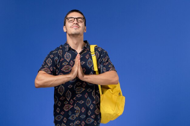Vista frontal del estudiante en camisa oscura con mochila amarilla posando en la pared azul