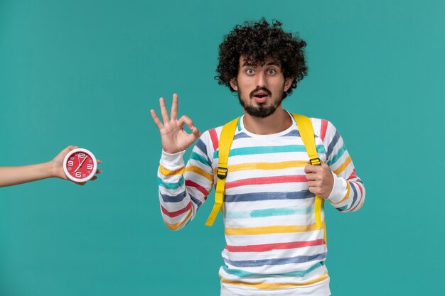 Vista frontal del estudiante en camisa a rayas con mochila amarilla posando en la pared azul claro