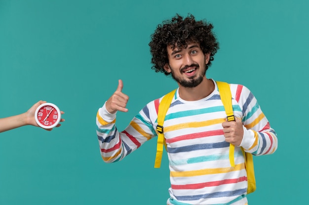 Vista frontal del estudiante en camisa a rayas con mochila amarilla posando con una sonrisa en la pared azul claro