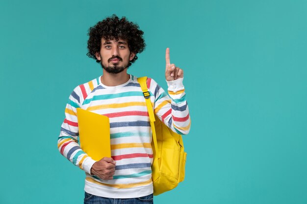 Vista frontal del estudiante en camisa a rayas con mochila amarilla sosteniendo archivos en la pared azul