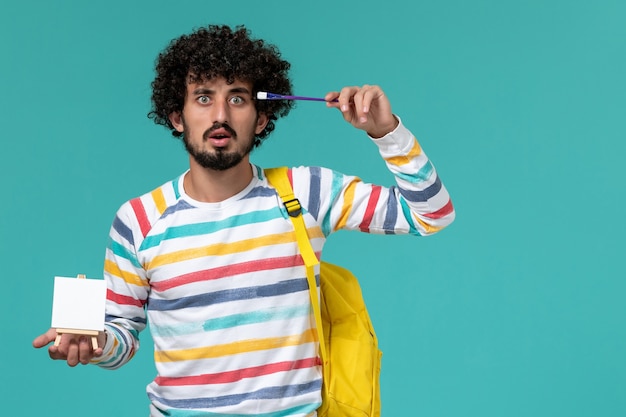 Vista frontal del estudiante en camisa a rayas con mochila amarilla sosteniendo caballete y borla en la pared azul