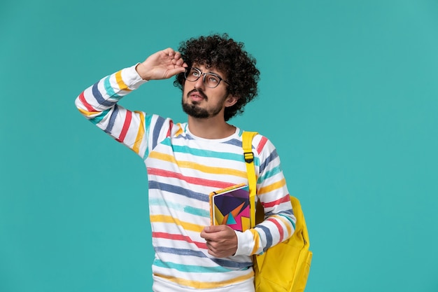 Vista frontal del estudiante en camisa a rayas con mochila amarilla sosteniendo cuadernos en la pared azul