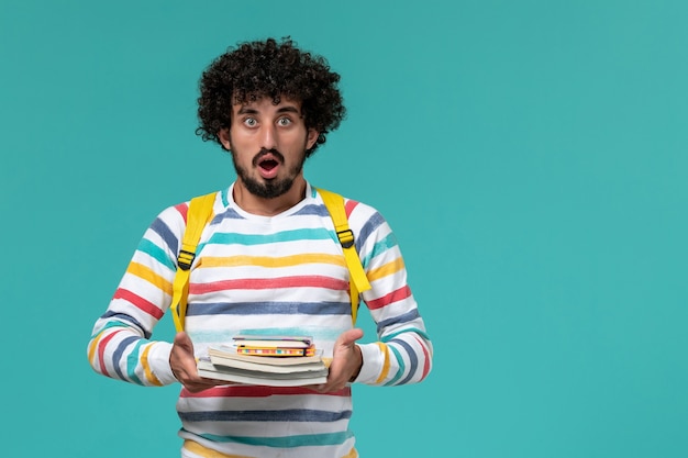 Vista frontal del estudiante en camisa a rayas con mochila amarilla sosteniendo libros en la pared azul