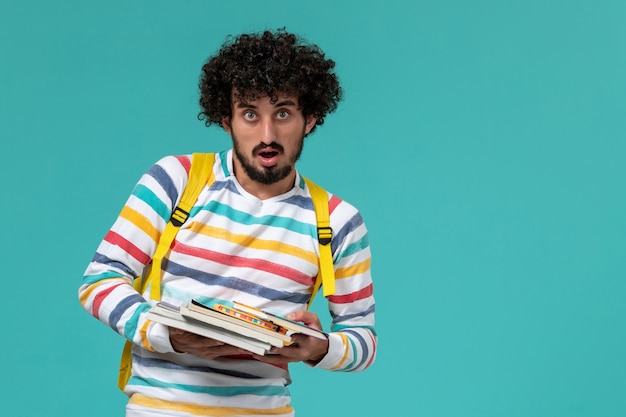 Vista frontal del estudiante en camisa a rayas con mochila amarilla sosteniendo libros en la pared azul