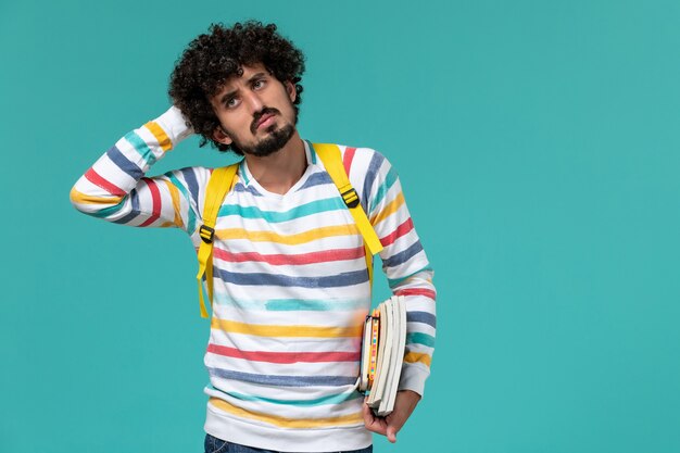 Vista frontal del estudiante en camisa a rayas con mochila amarilla sosteniendo libros y pensando en la pared azul