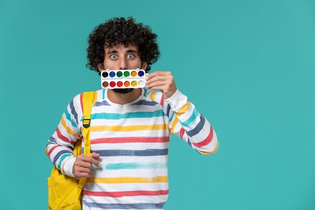 Vista frontal del estudiante en camisa a rayas con mochila amarilla sosteniendo pinturas en la pared azul