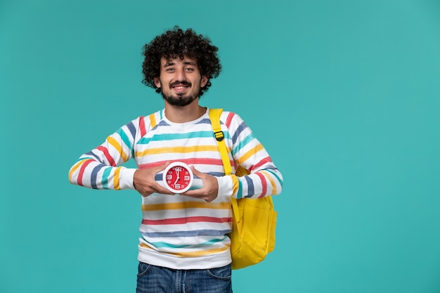 Vista frontal del estudiante en camisa a rayas con mochila amarilla sosteniendo relojes en la pared azul