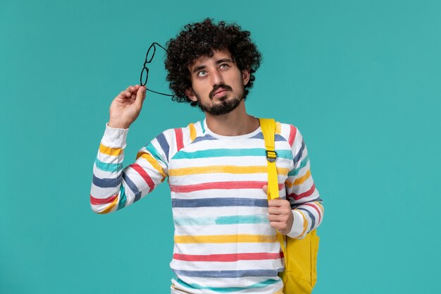 Vista frontal del estudiante en camisa a rayas con mochila amarilla sosteniendo sus gafas de sol ópticas pensando en la pared azul