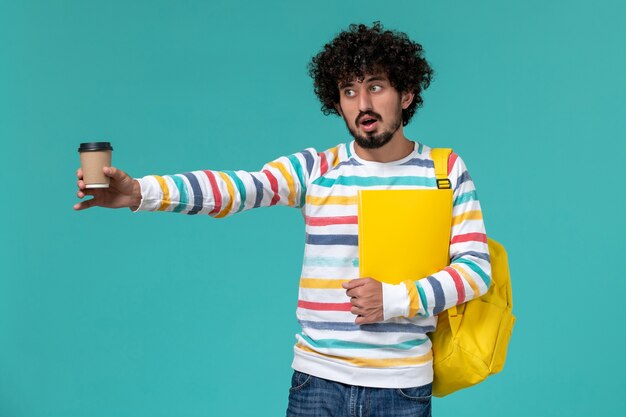 Vista frontal del estudiante en camisa a rayas vistiendo una mochila amarilla sosteniendo archivos y café en la pared azul
