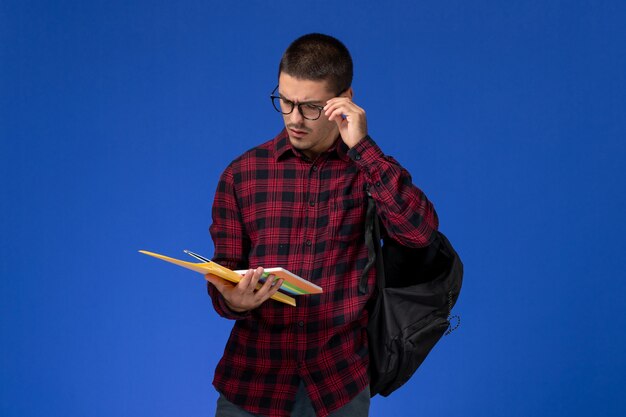 Vista frontal del estudiante en camisa roja a cuadros con mochila con archivos y cuaderno en la pared azul