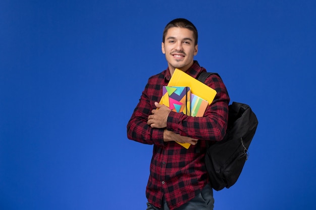Vista frontal del estudiante en camisa roja a cuadros con mochila con cuaderno y archivos en la pared azul claro