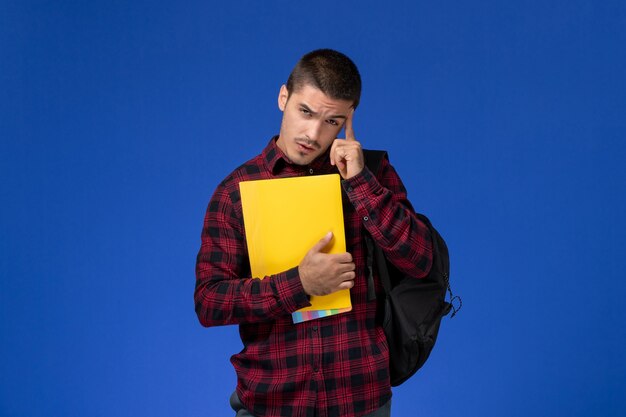 Vista frontal del estudiante en camisa roja a cuadros con mochila sosteniendo archivos amarillos en la pared azul