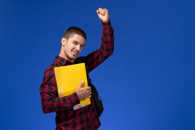 Vista frontal del estudiante en camisa roja a cuadros con mochila sosteniendo archivos amarillos en la pared azul