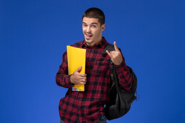 Foto gratuita vista frontal del estudiante en camisa roja a cuadros con mochila sosteniendo archivos amarillos en la pared azul