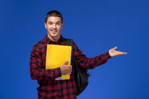 Vista frontal del estudiante en camisa roja a cuadros con mochila sosteniendo archivos amarillos en la pared azul