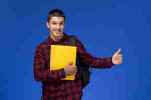 Foto gratuita vista frontal del estudiante en camisa roja a cuadros con mochila sosteniendo archivos amarillos en la pared azul