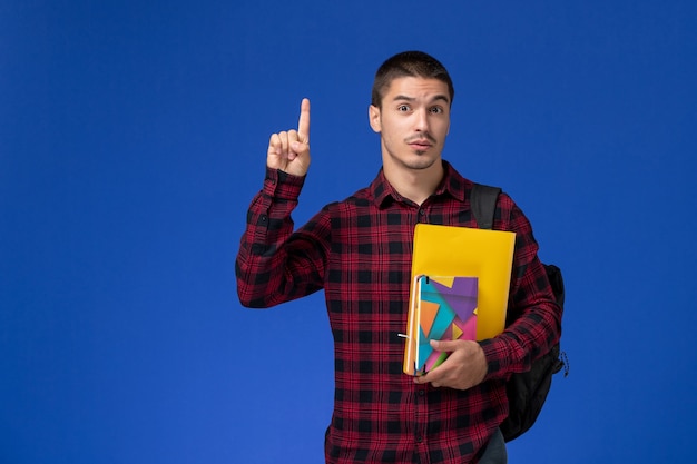 Vista frontal del estudiante en camisa roja a cuadros con mochila sosteniendo archivos y cuadernos levantando el dedo sobre la pared azul