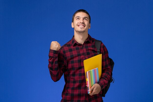 Vista frontal del estudiante en camisa roja a cuadros con mochila sosteniendo archivos y cuadernos regocijándose en la pared azul