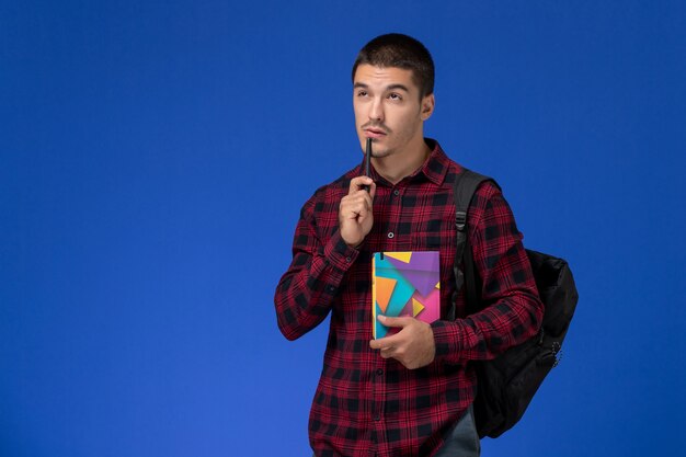 Vista frontal del estudiante en camisa roja a cuadros con mochila sosteniendo el cuaderno pensando en la pared azul claro