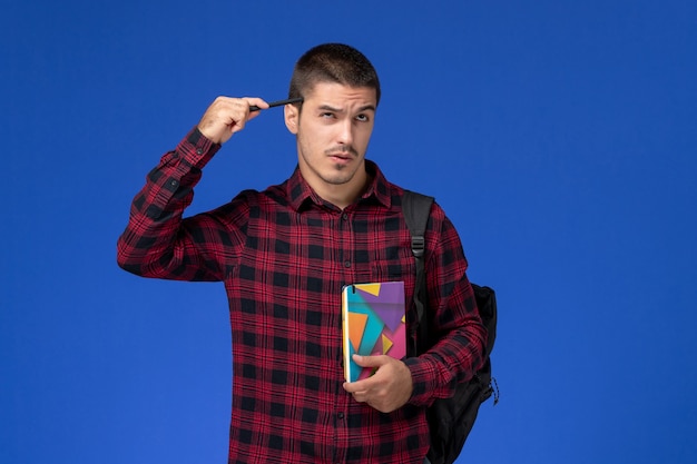 Foto gratuita vista frontal del estudiante en camisa roja a cuadros con mochila sosteniendo el cuaderno pensando en la pared azul claro