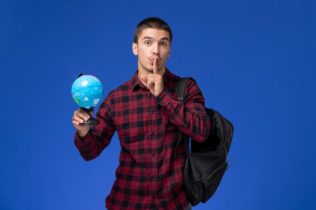 Vista frontal del estudiante en camisa roja a cuadros con mochila sosteniendo un pequeño globo en la pared azul claro