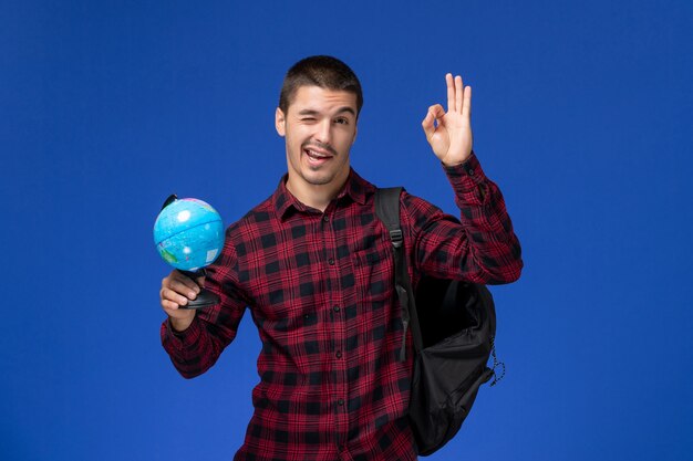 Vista frontal del estudiante en camisa roja a cuadros con mochila sosteniendo un pequeño globo en la pared azul claro