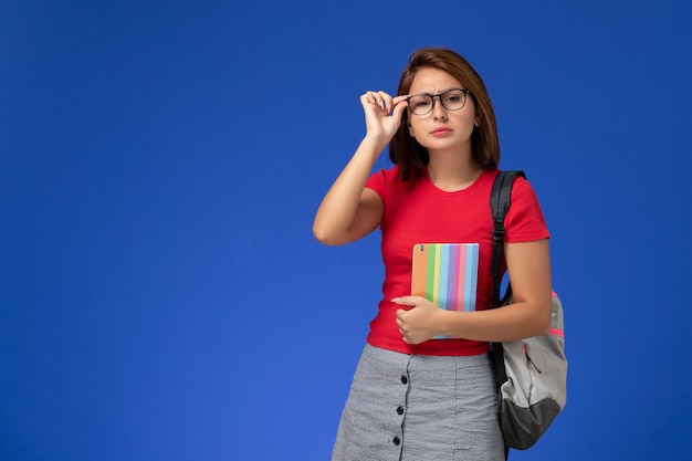 Vista frontal de la estudiante en camisa roja con mochila con cuaderno en la pared azul