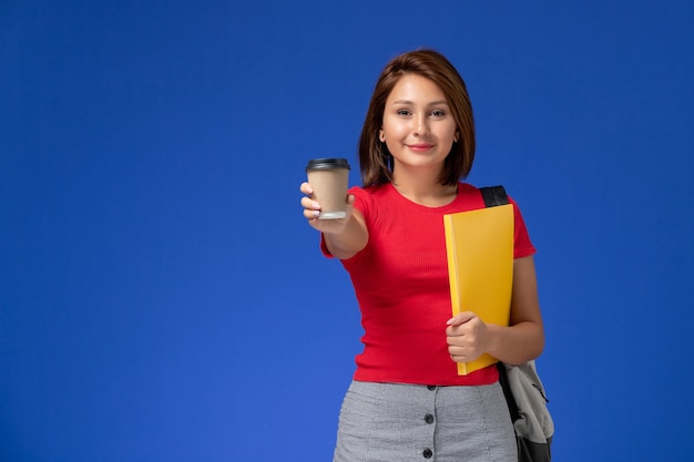 Vista frontal de la estudiante en camisa roja con mochila sosteniendo archivos amarillos y café en la pared azul