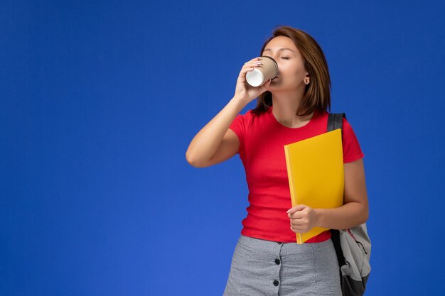 Vista frontal de la estudiante en camisa roja con mochila sosteniendo archivos amarillos tomando café en la pared azul