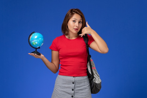 Vista frontal de la estudiante en camisa roja con mochila sosteniendo globo pequeño posando en la pared azul