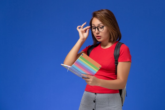 Vista frontal de la estudiante en camisa roja con mochila sosteniendo la lectura del cuaderno en la pared azul