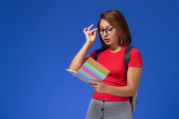 Vista frontal de la estudiante en camisa roja con mochila sosteniendo la lectura del cuaderno en la pared azul