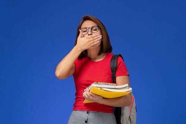 Vista frontal de la estudiante en camisa roja con mochila sosteniendo libros y archivos bostezando en la pared azul