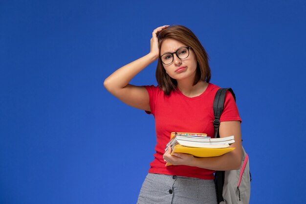 Vista frontal de la estudiante en camisa roja con mochila sosteniendo libros y archivos deprimidos en la pared azul claro