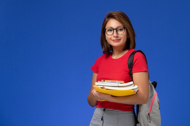 Vista frontal de la estudiante en camisa roja con mochila sosteniendo libros y archivos sonriendo en la pared azul