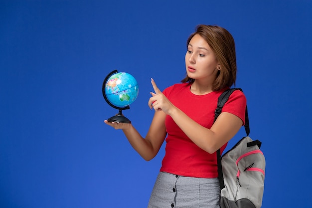 Vista frontal de la estudiante en camisa roja con mochila sosteniendo un pequeño globo redondo en la pared azul