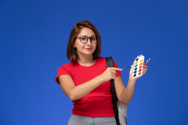 Vista frontal de la estudiante en camisa roja con mochila sosteniendo pinturas para dibujar y borlas sonriendo en la pared azul