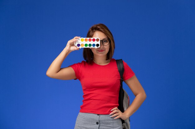 Vista frontal de la estudiante en camisa roja con mochila sosteniendo pinturas para dibujar