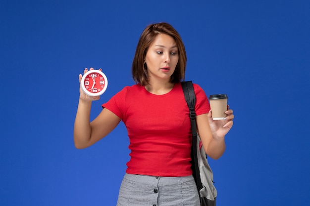 Vista frontal de la estudiante en camisa roja con mochila sosteniendo relojes y café en la pared azul