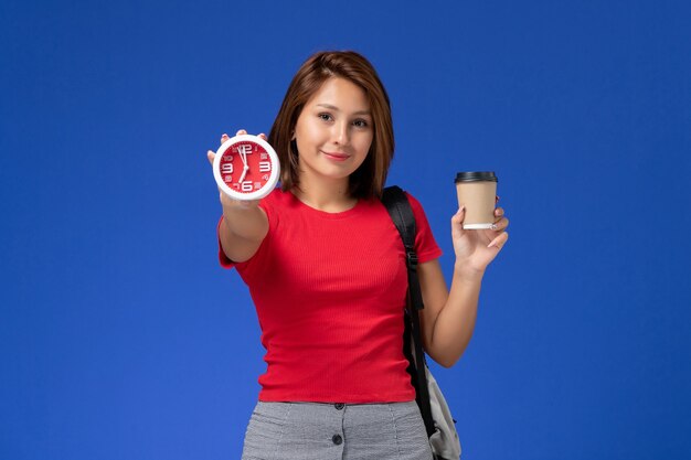 Vista frontal de la estudiante en camisa roja con mochila sosteniendo relojes y café en la pared azul