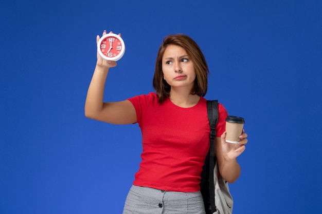 Vista frontal de la estudiante en camisa roja con mochila sosteniendo relojes y café en la pared azul