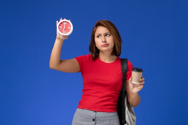 Vista frontal de la estudiante en camisa roja con mochila sosteniendo relojes y café en la pared azul