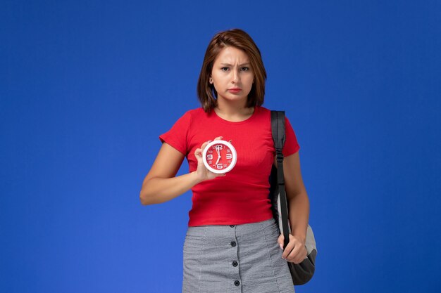 Vista frontal de la estudiante en camisa roja con mochila sosteniendo relojes en la pared azul